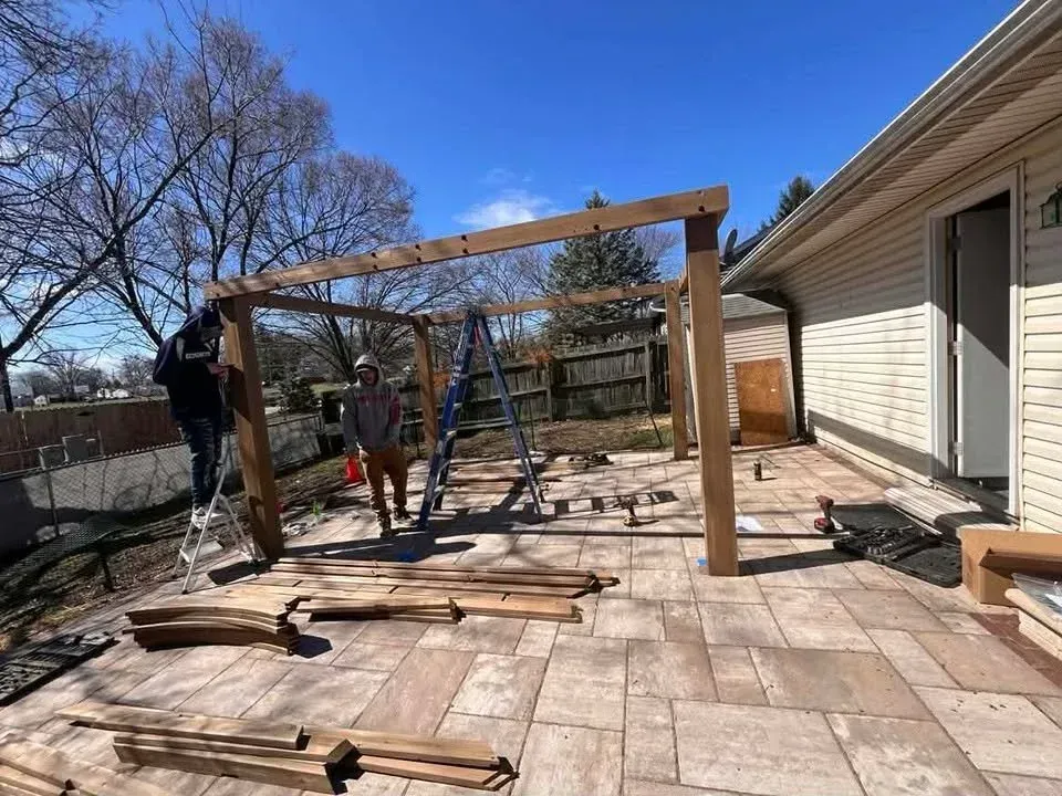 Workers assembling a wooden pergola frame on a sunlit backyard patio beside a house.