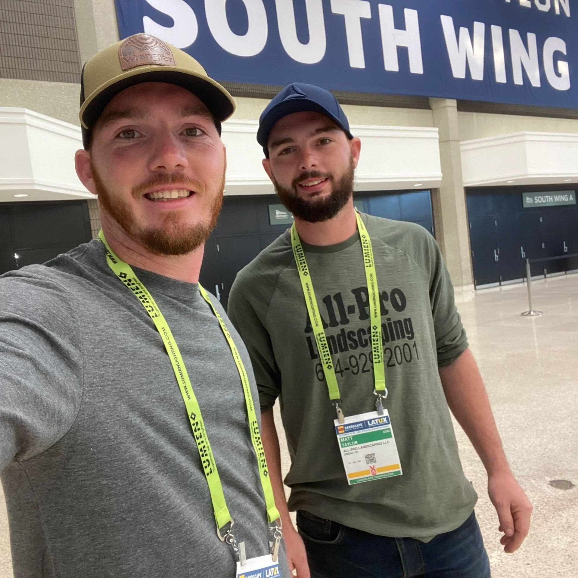 Two smiling attendees wearing lanyards pose indoors beneath a “South Wing” sign.
