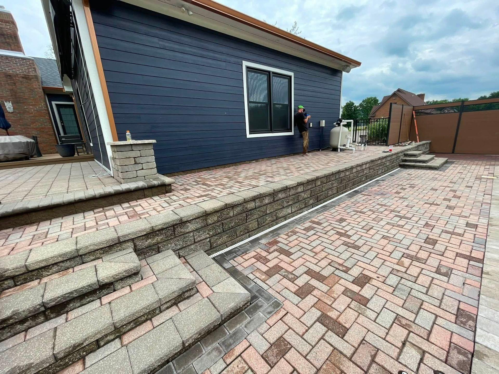 Brick driveway and steps beside a dark blue house with a paved patio and two people near the side entrance