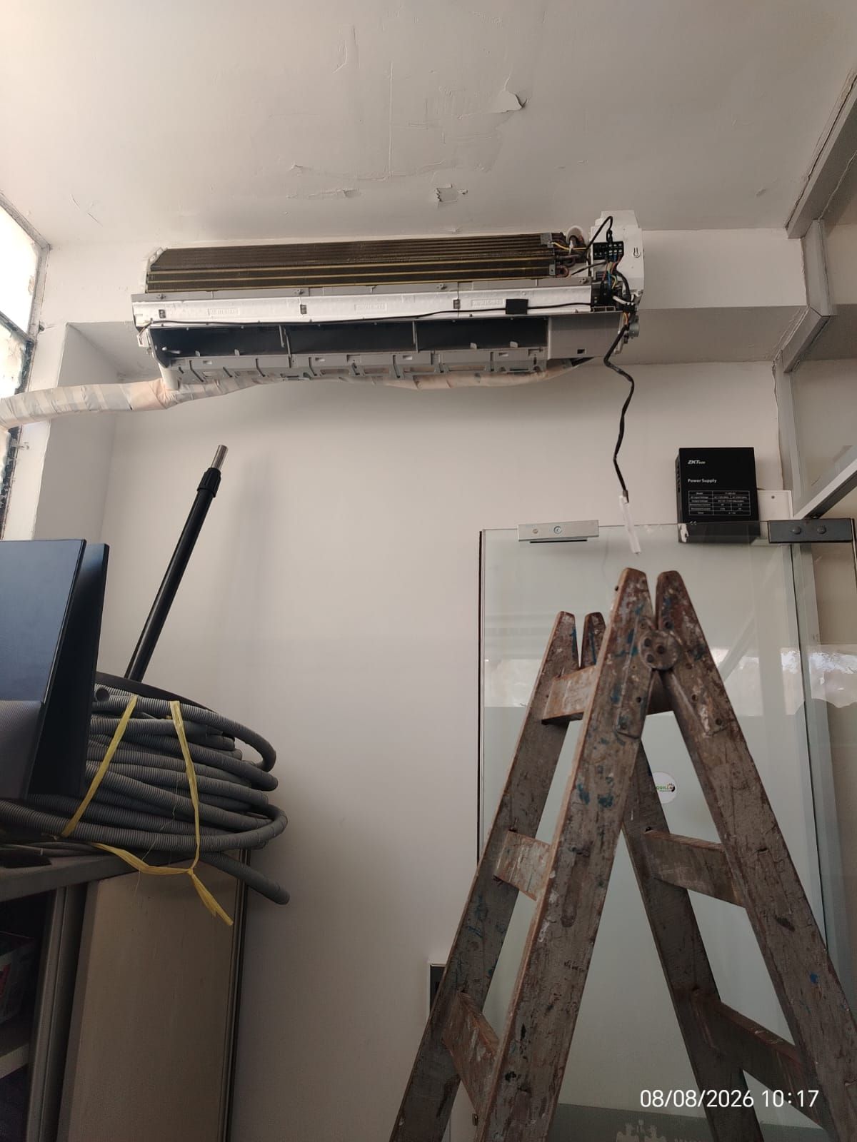Aire acondicionado de pared parcialmente desmontado sobre escaleras y artículos de limpieza en una habitación.
