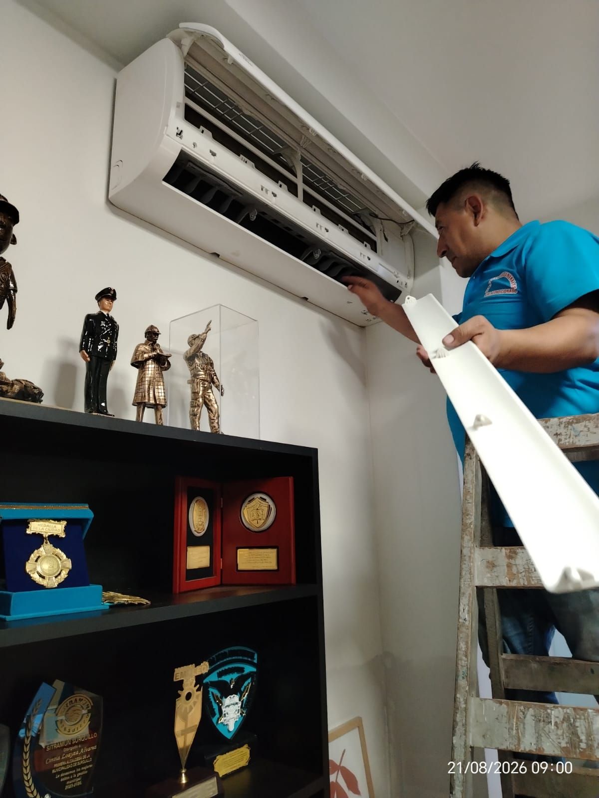 Técnico reparando un aire acondicionado de pared situado encima de una estantería con trofeos y figuritas.