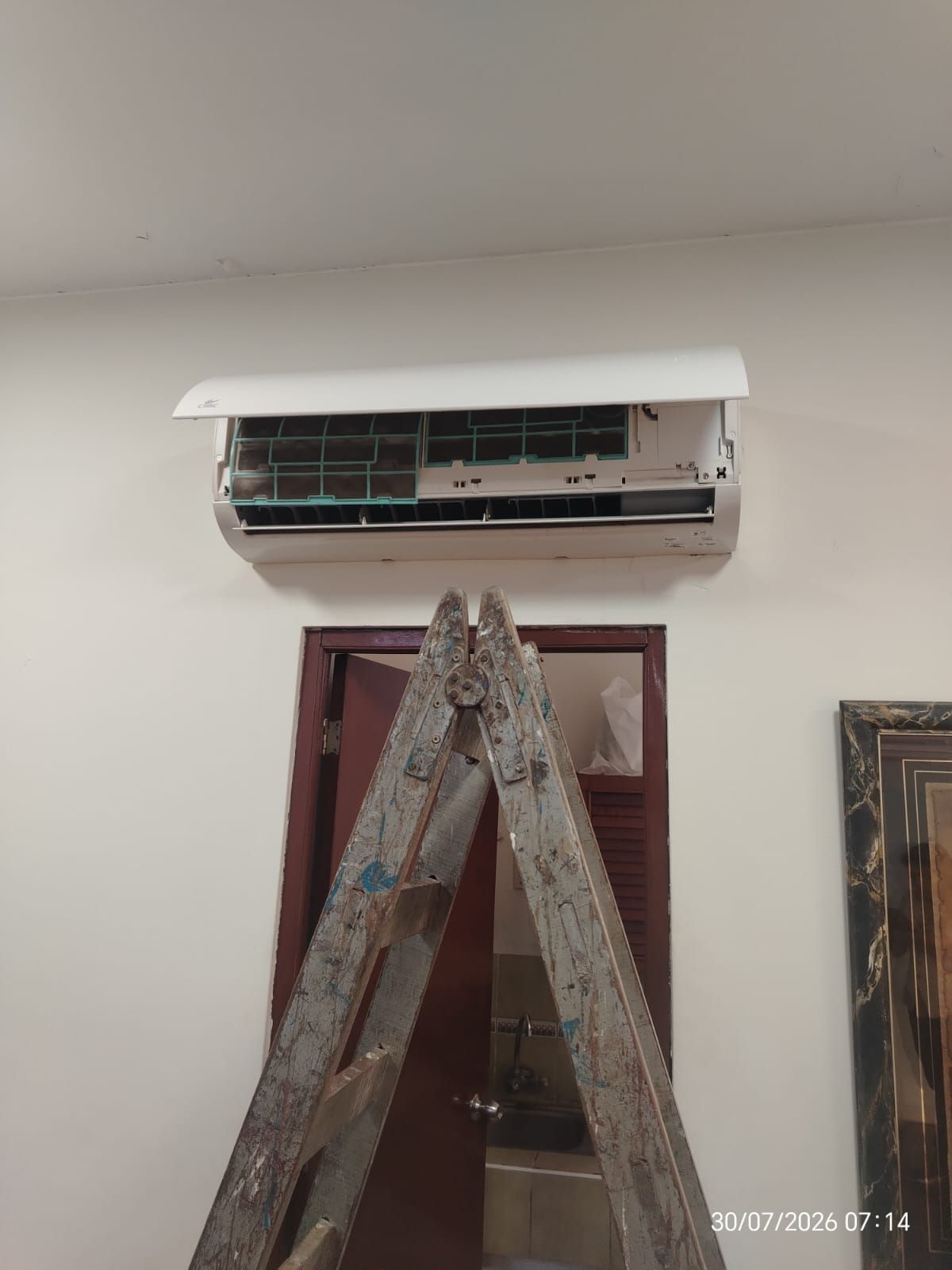 Aire acondicionado mural parcialmente instalado sobre una puerta, con una escalera apoyada debajo.