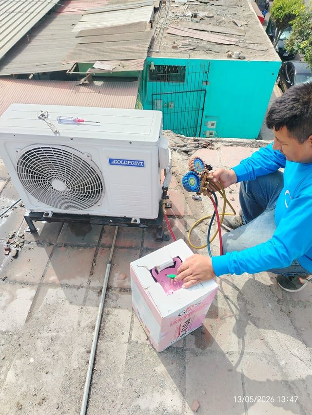 Técnico vestido de azul trabajando en una unidad de aire acondicionado en la azotea con un manómetro y herramientas.