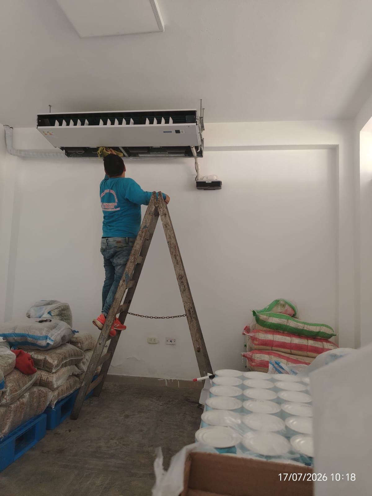 Un trabajador subido a una escalera instala un aire acondicionado de techo en una habitación blanca con materiales apilados.