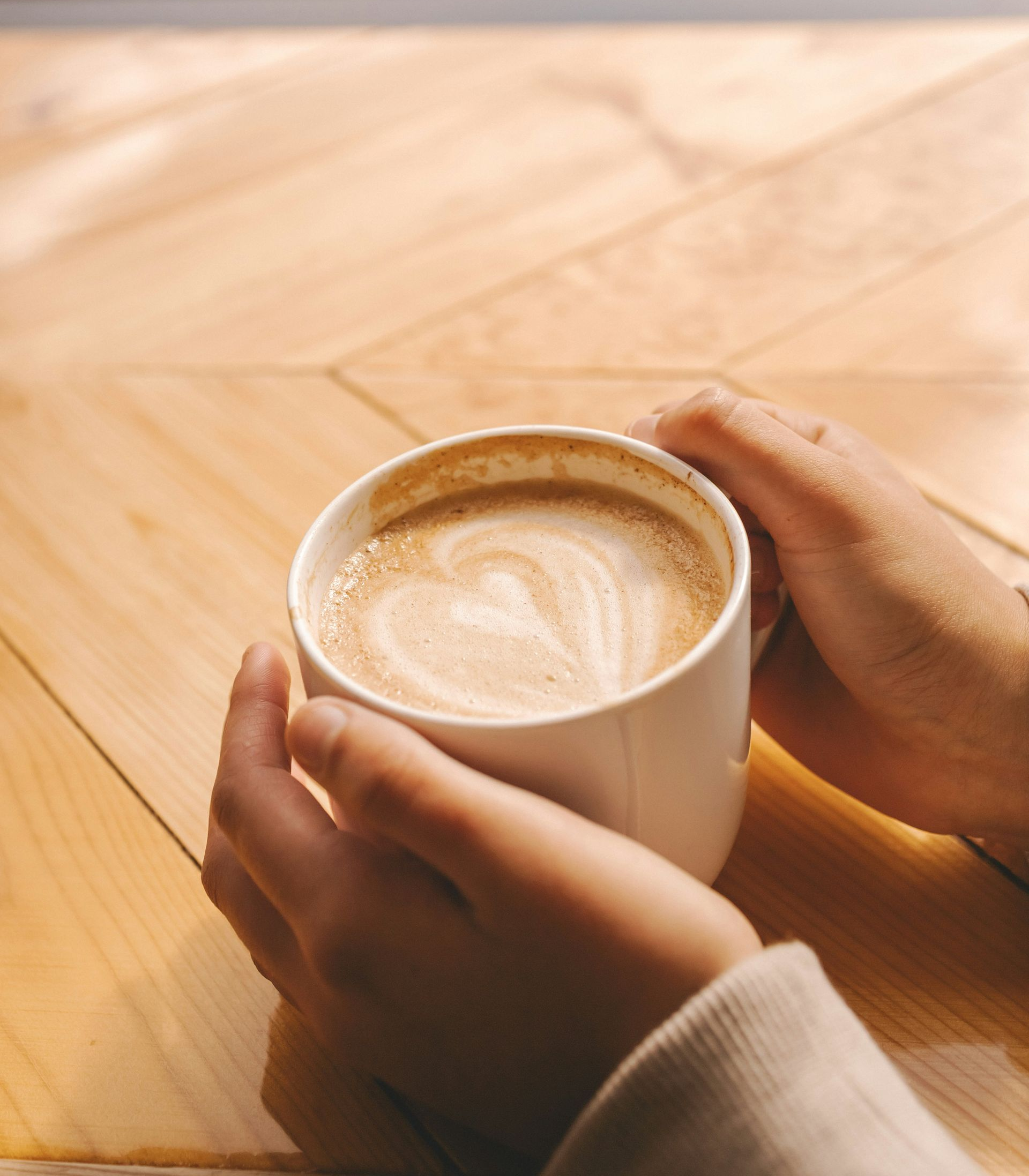 Hands holding a white mug of latte on a wooden table.