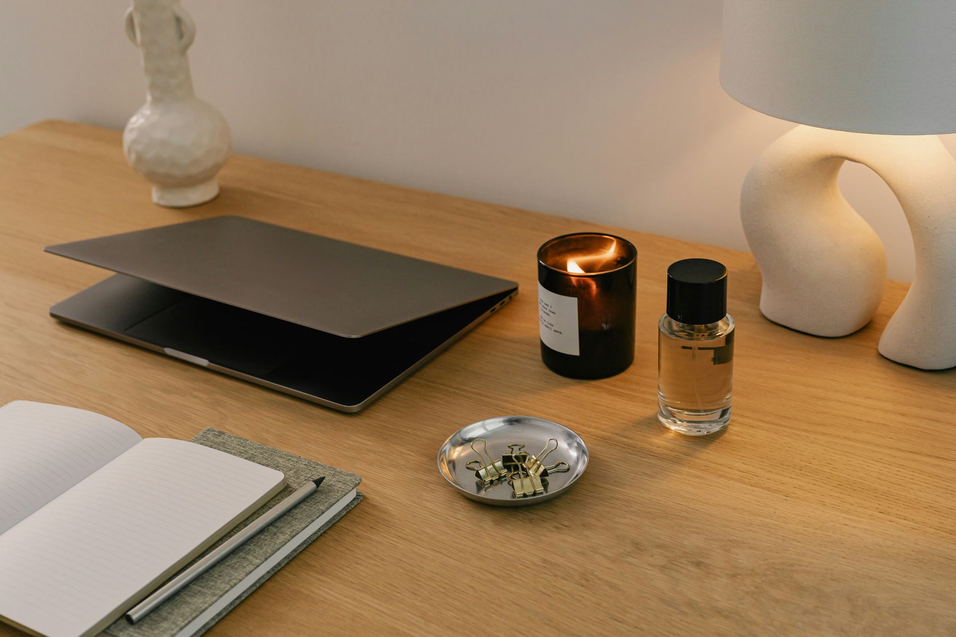 Desk with open notebook, closed laptop, candle, perfume bottle, and small dish on a wooden surface.