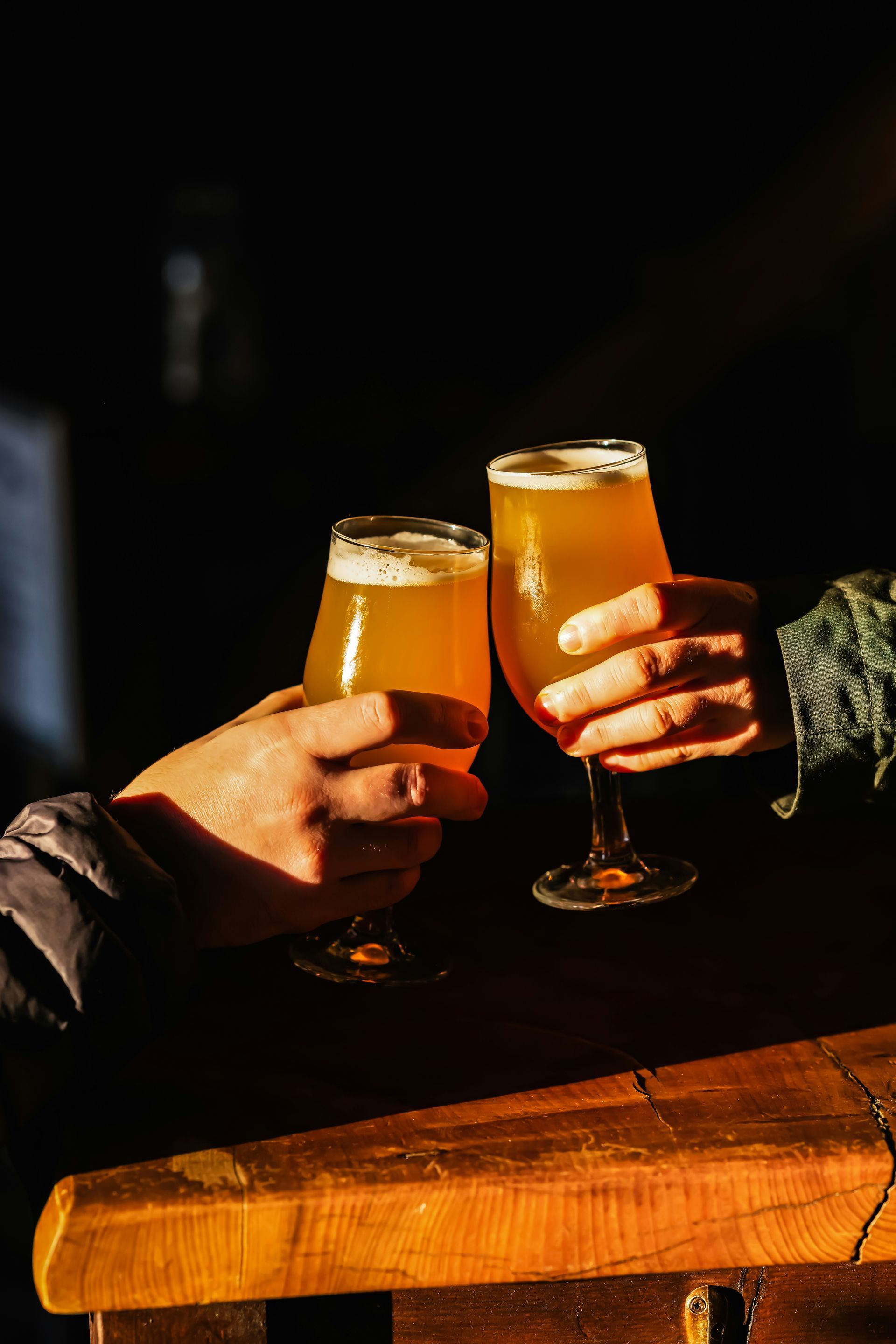 Two hands clink tall beer glasses over a wooden table in warm light.