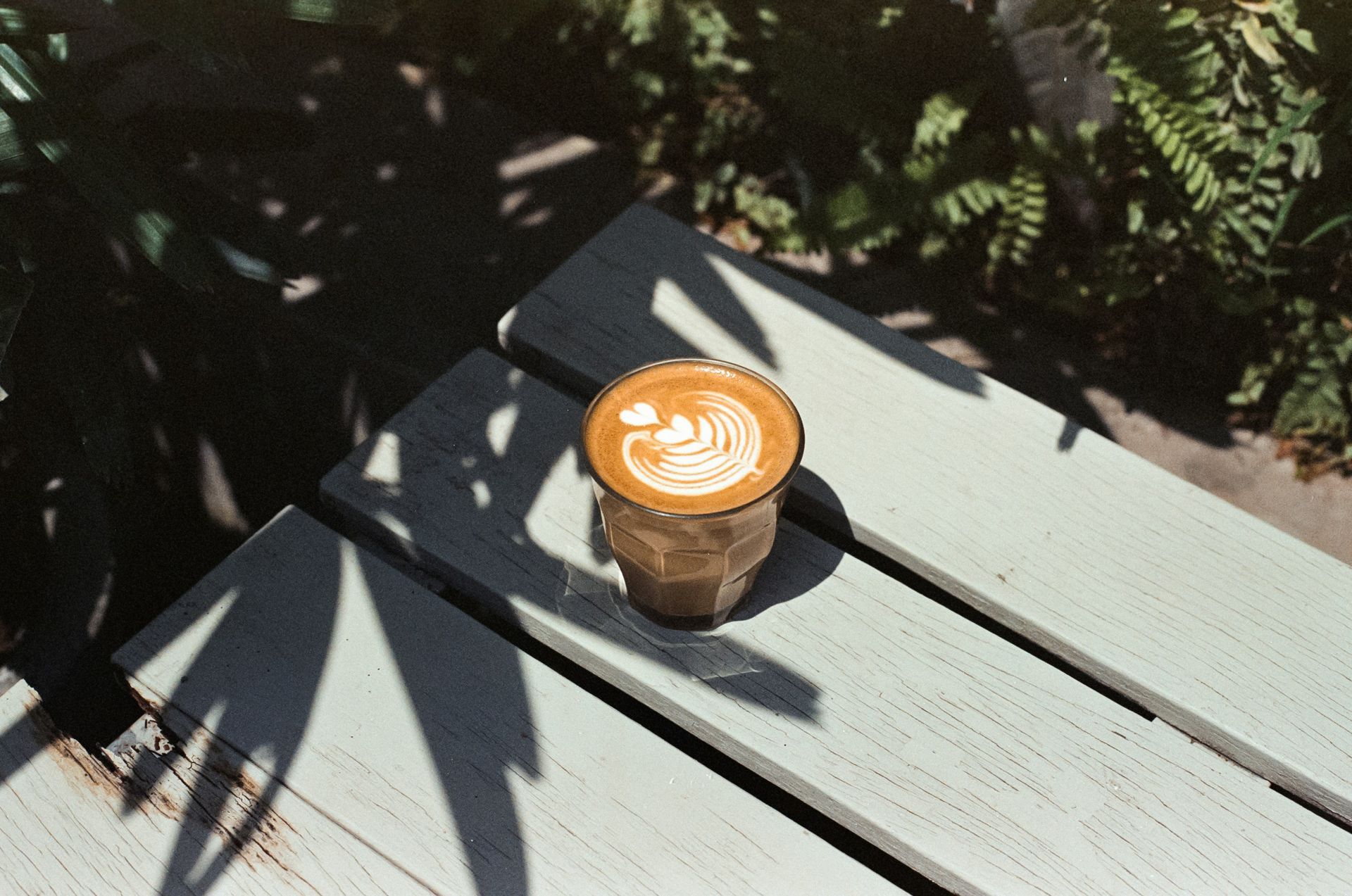 Iced latte in a clear cup on a sunlit gray bench outdoors, with plant shadows nearby
