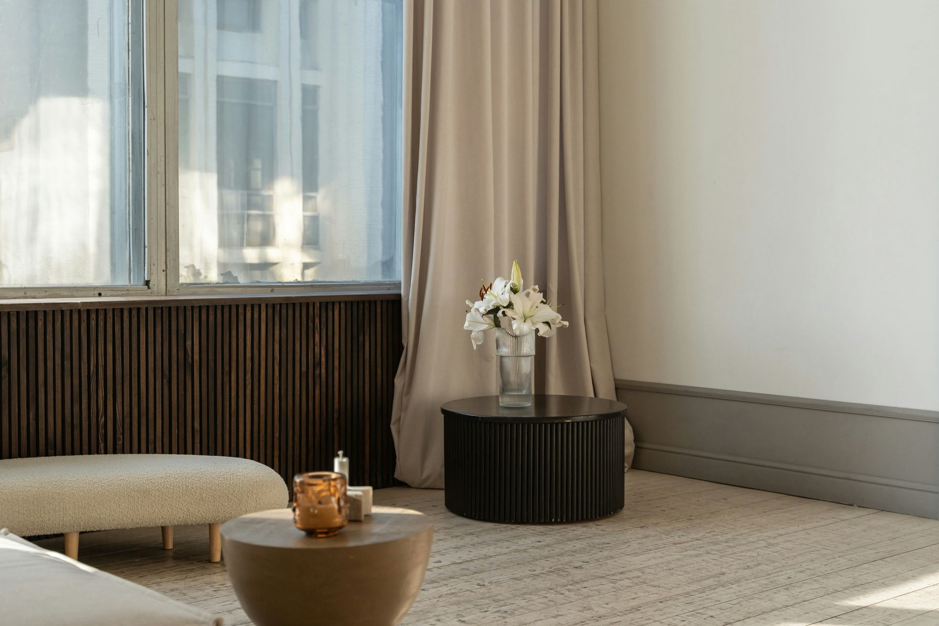 Modern living room with large windows, beige curtains, dark wood paneling, and a small round table with flowers.