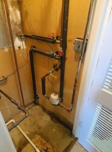 Narrow utility closet with exposed pipes, valves, and a small white water meter on a yellow wall.