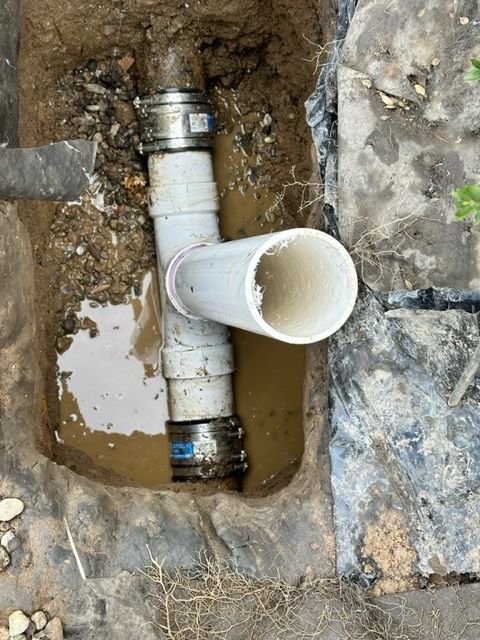 Open white PVC drain pipe in a rocky trench with metal couplings.
