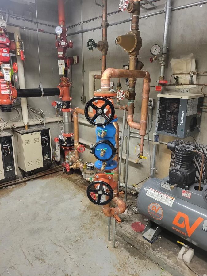 Industrial mechanical room with pipes, valves, and pumps, including a large gray air compressor on the right.