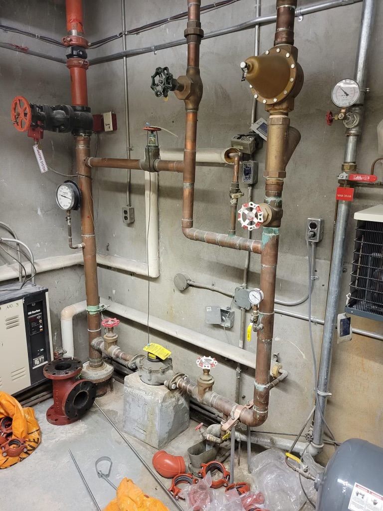 Mechanical room with copper pipes, valves, gauges, and a boiler on the floor near a wall-mounted unit.