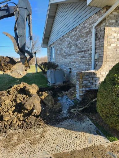Excavator digging beside a brick house, with dirt piled by the foundation and a muddy trench.
