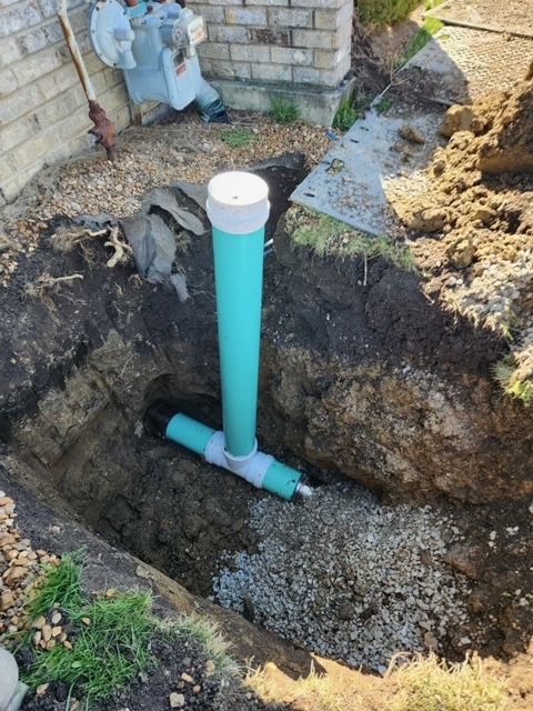 Excavated hole exposing a turquoise PVC pipe and white cap beside a brick wall and dirt pile