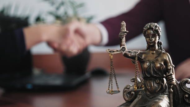 Small Lady Justice statue in foreground with blurred handshake and laptop in the background.
