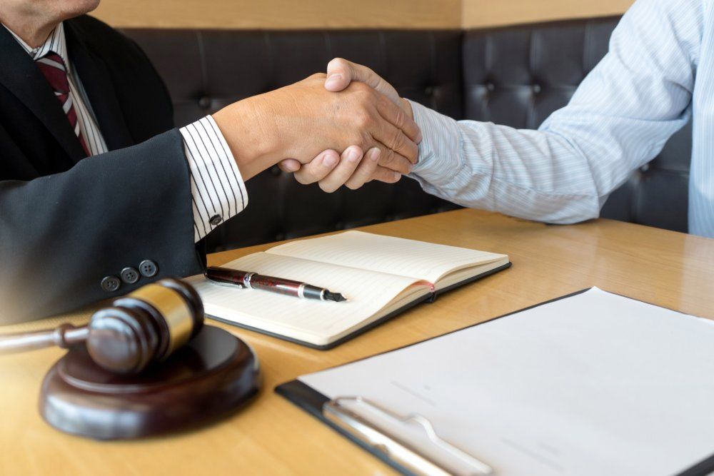 Two people shake hands across a desk with a gavel, notebook, and papers in a legal office.