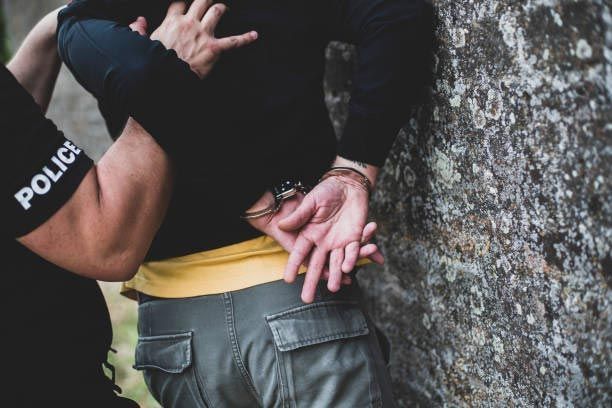 Police officer handcuffing a person against a stone wall, close-up of restrained hands.