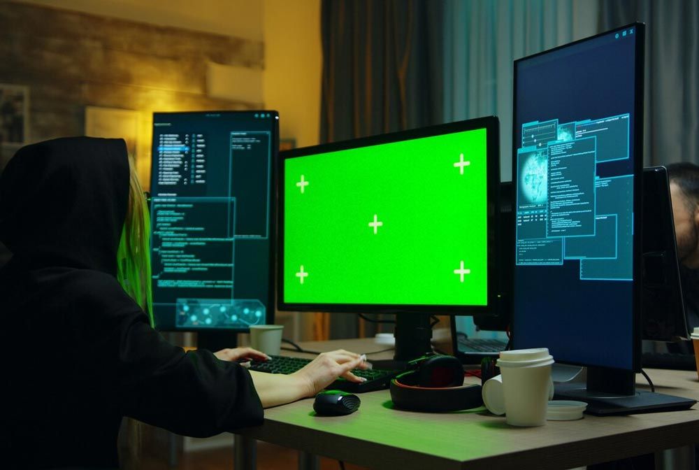 Hooded person working at a desk with multiple monitors, including a green screen, in a dimly lit room