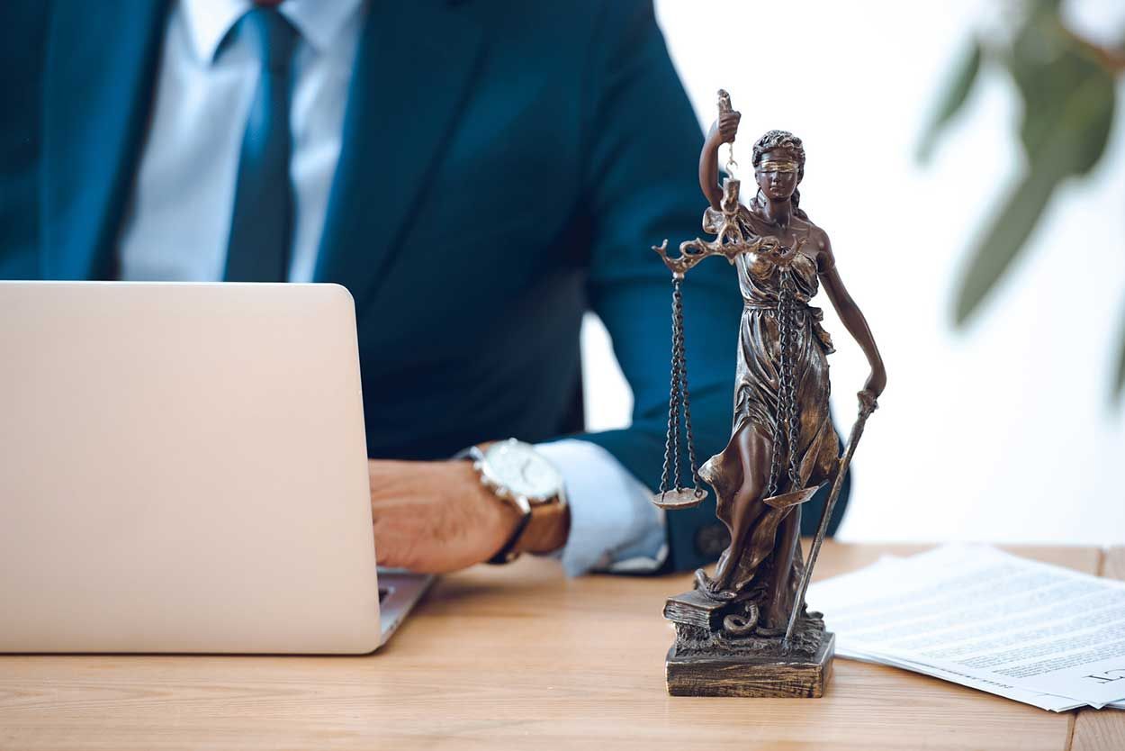 Desk with laptop, businessperson, and Lady Justice statue beside legal papers