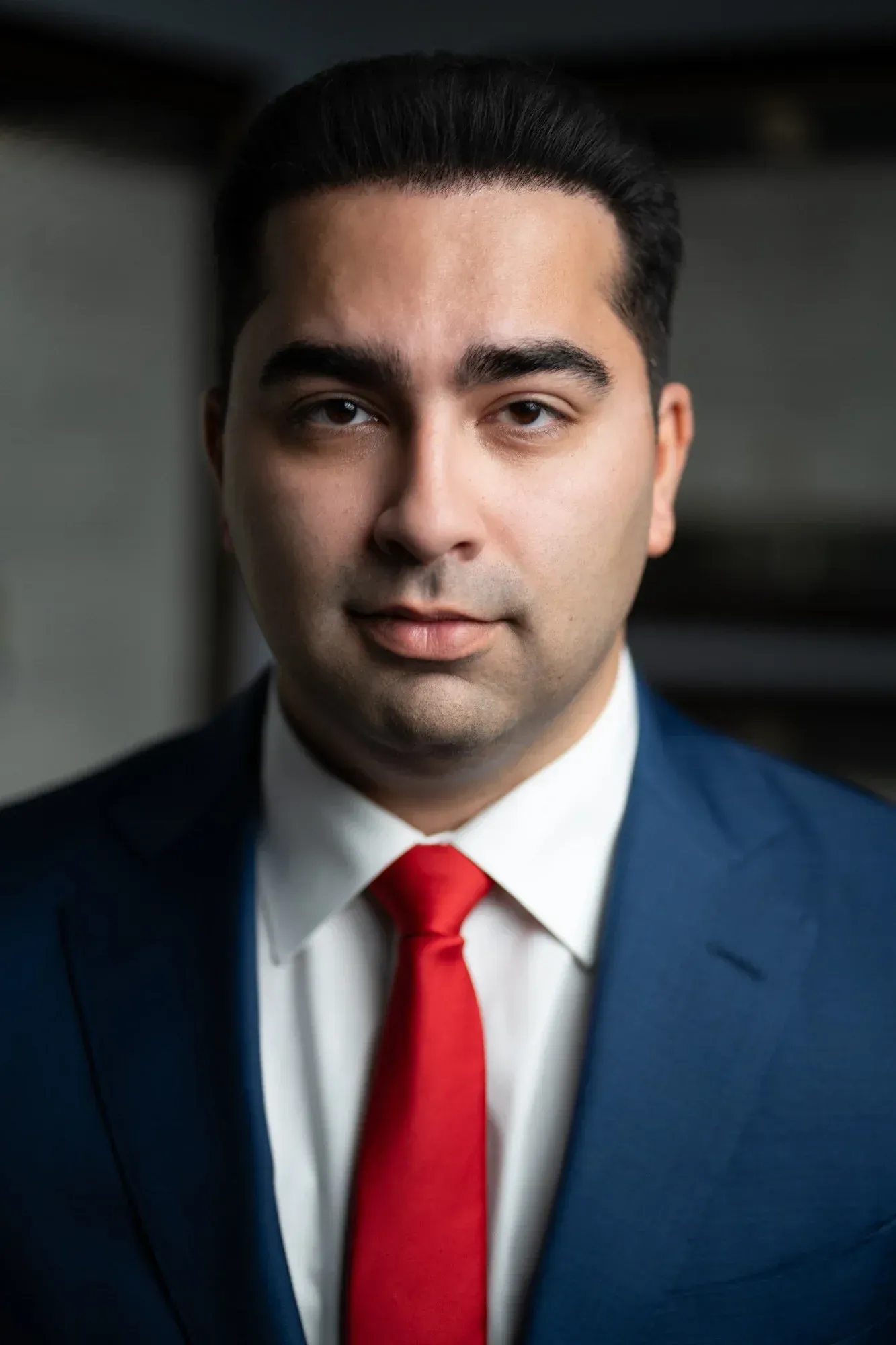 Formal headshot of a man in a navy suit and red tie against a dark blurred background