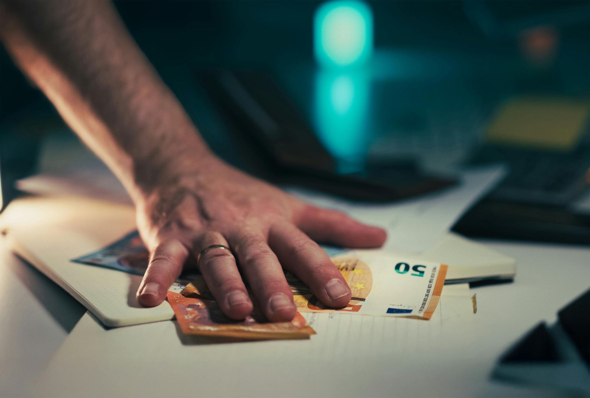 Hand placing cash on a desk beside a 50-euro note and laptop, with a blurred teal background