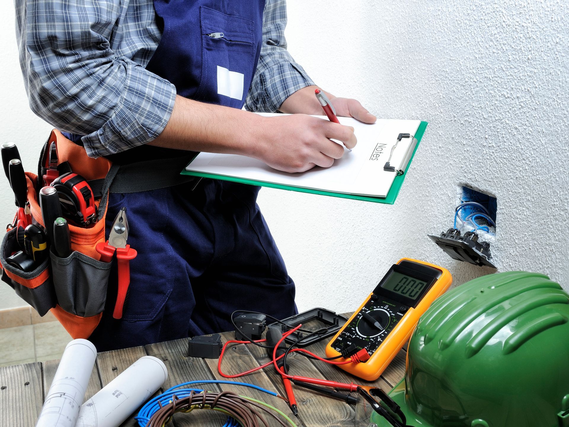 Electrician in overalls writing on clipboard, examining electrical equipment in a workshop.