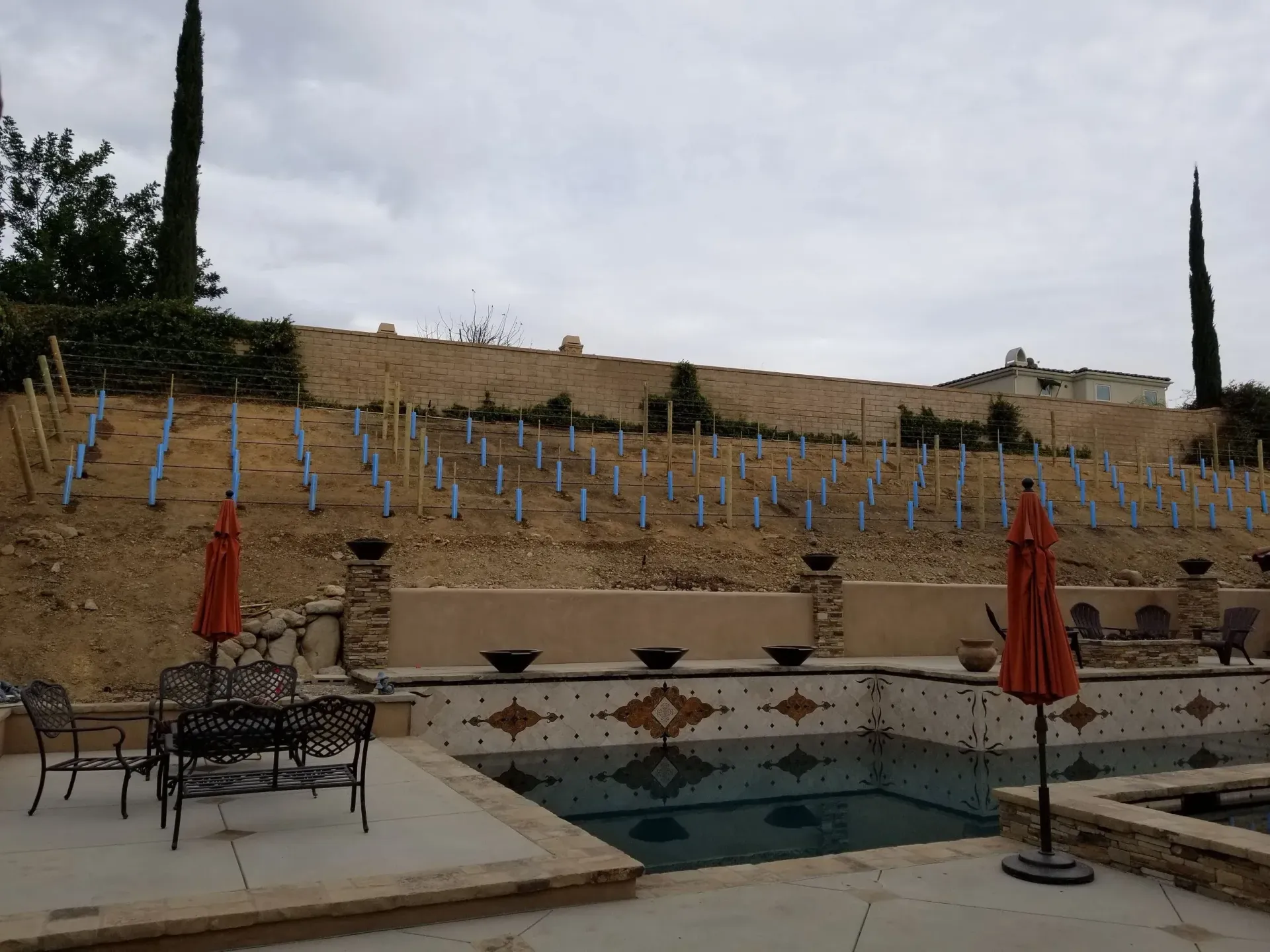Pool area with umbrellas and furniture, hillside vineyard in the background under a cloudy sky.
