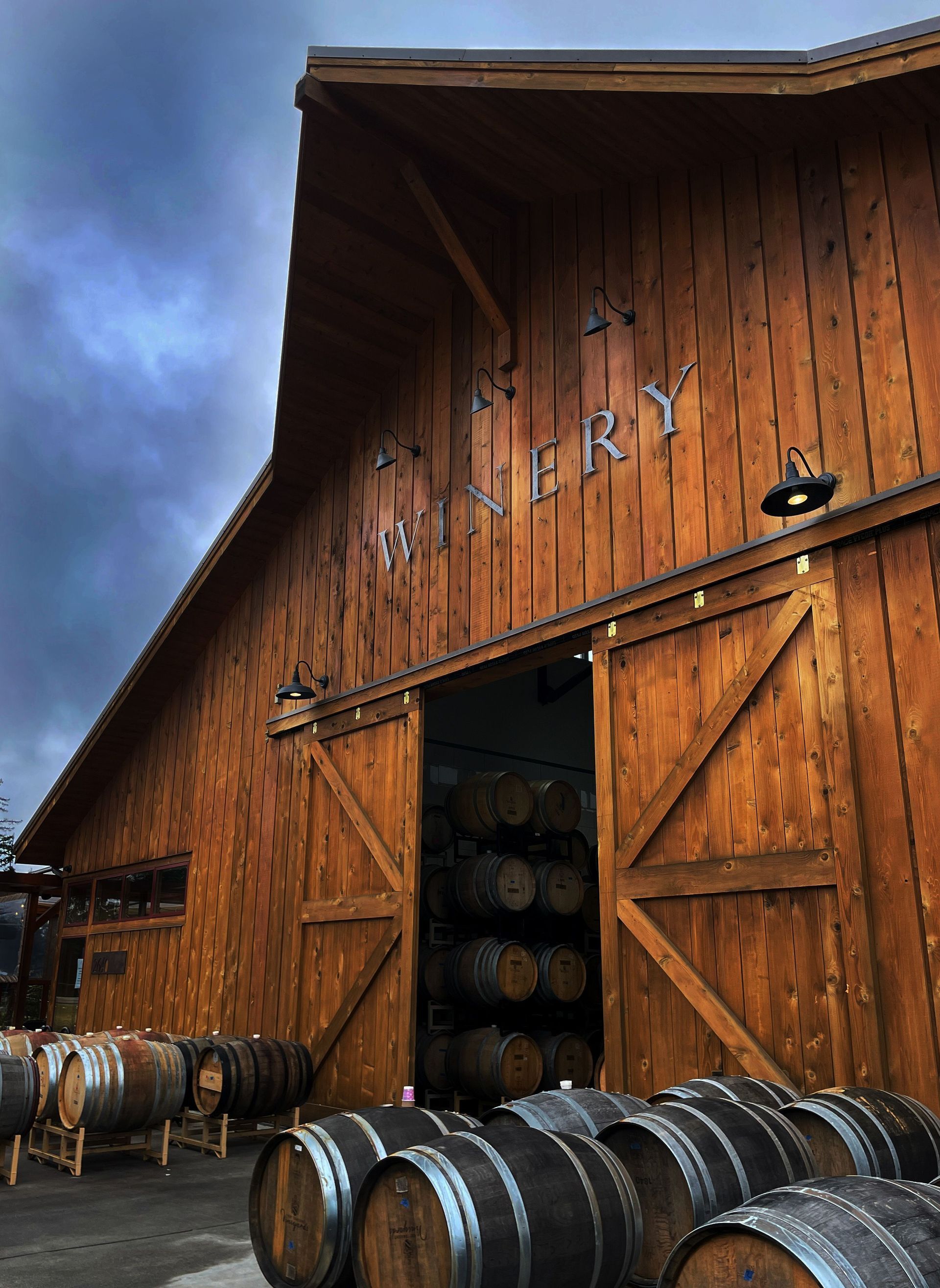 Wooden winery barn with open doors and stacked wine barrels outside under a cloudy sky