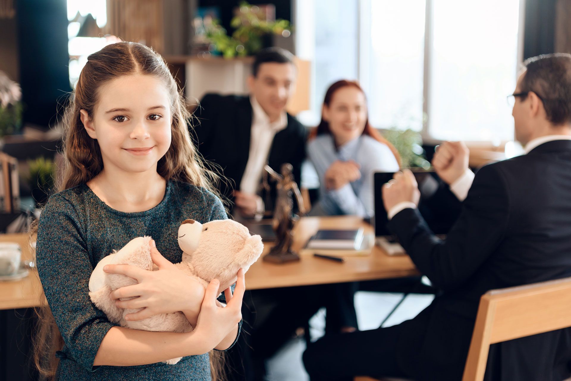 A little girl is holding a teddy bear in front of a family sitting at a table