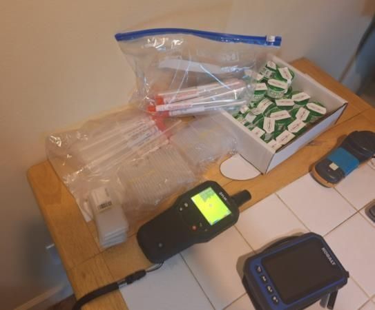 Testing supplies, including swabs and small vials in plastic bags and a box, alongside two handheld electronic devices.