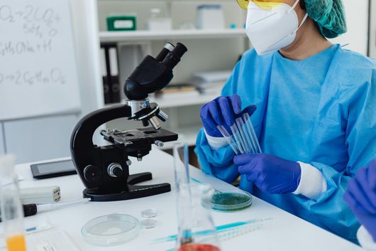 A scientist in a blue gown, mask, and gloves holding test tubes at a lab desk with a microscope.