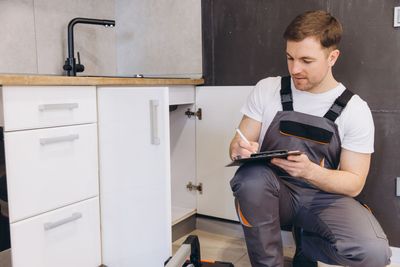 A person in a gray work jumpsuit kneels on the floor, writing on a clipboard while inspecting an open kitchen cabinet.