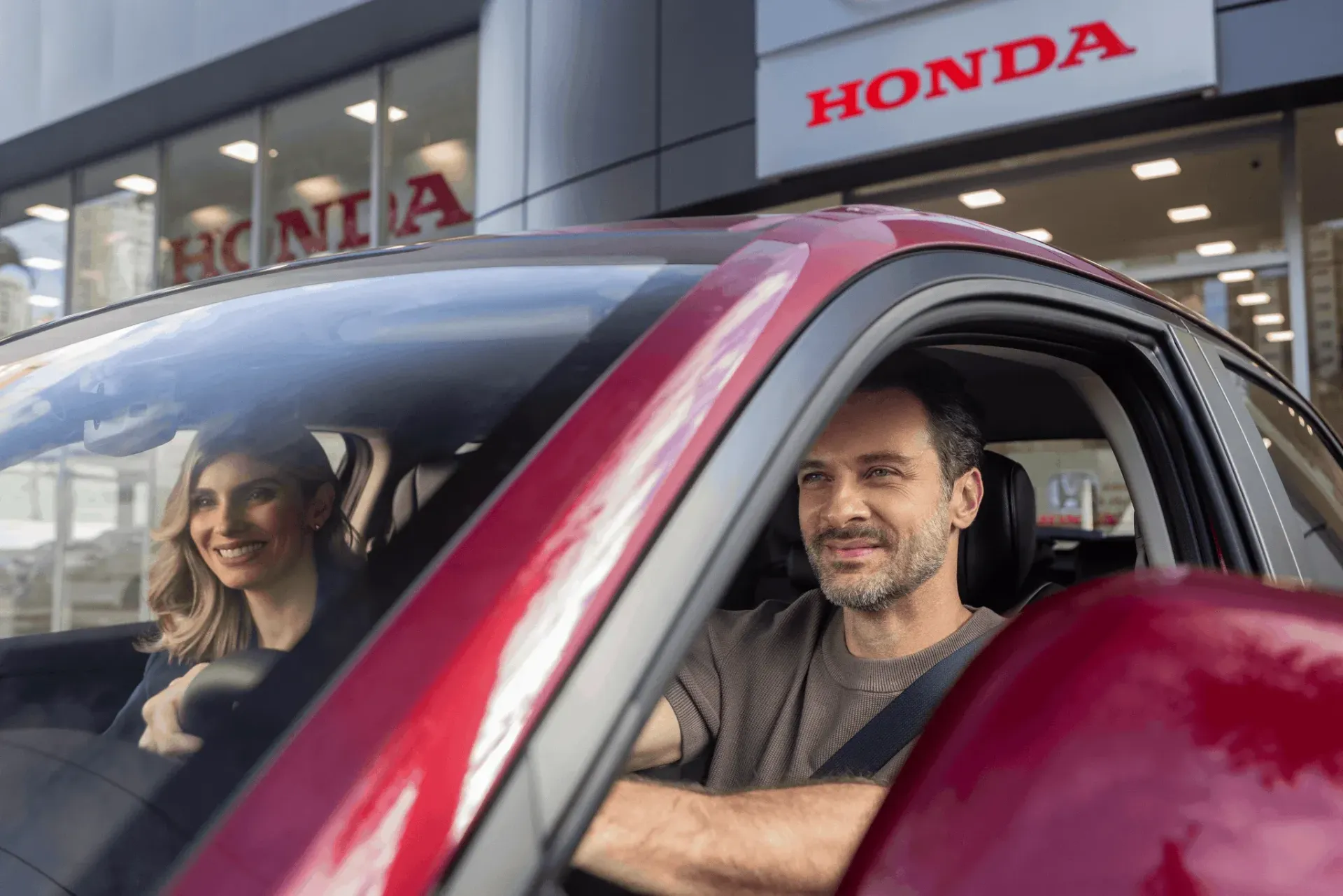 Duas pessoas estão sentadas em um carro vermelho em frente a uma concessionária Honda, sorrindo para a câmera.