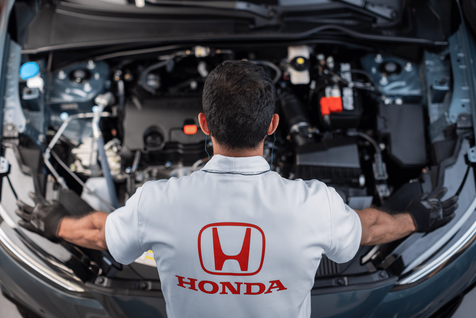 Mecânico da Honda trabalhando sob o capô de um carro em uma oficina, vestindo uma camisa branca da Honda.