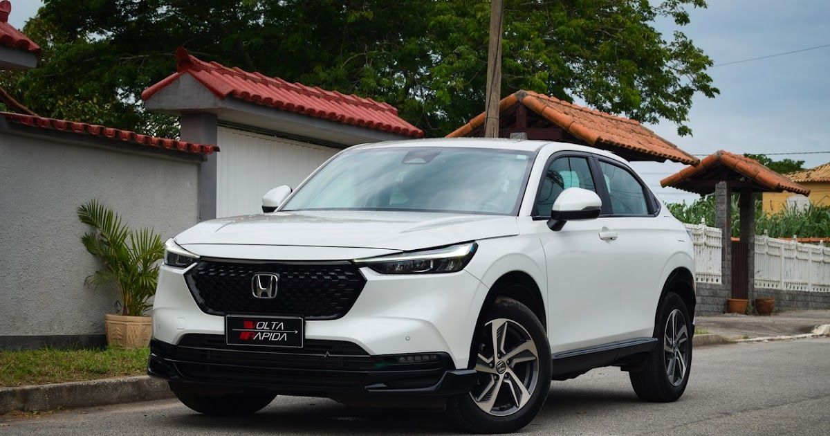 SUV branca estacionada em uma rua molhada ao lado de um muro cinza e árvores.