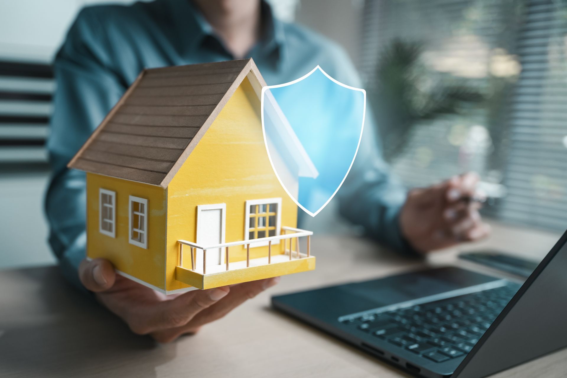 Yellow house model with blue shield in front of a person at a laptop, suggesting home insurance