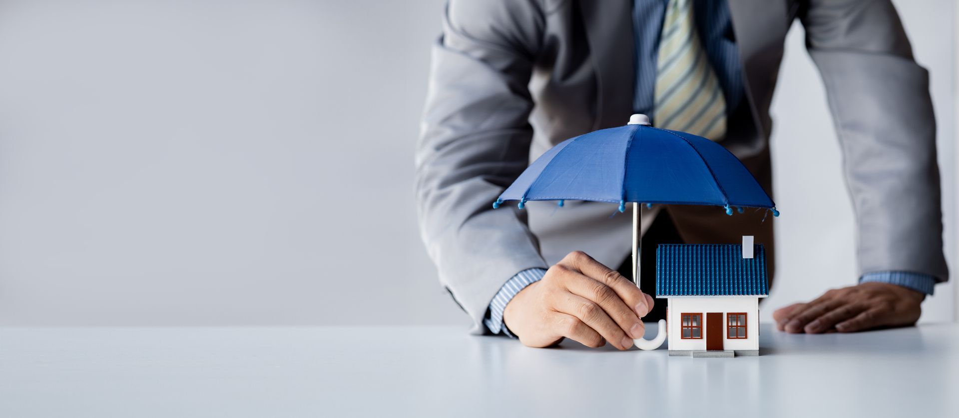 A person in a gray suit places a small house model under a blue umbrella, symbolizing home insurance.