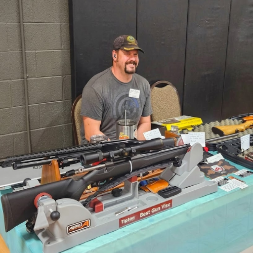 Man seated behind a table displaying several scoped rifles and firearm accessories at a gun show
