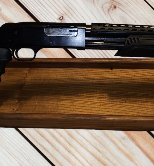 Close-up of a black shotgun resting on a wooden rack, with the receiver and barrel visible.