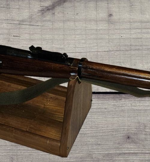 Wooden-stock rifle resting on a wooden display stand against a light background.