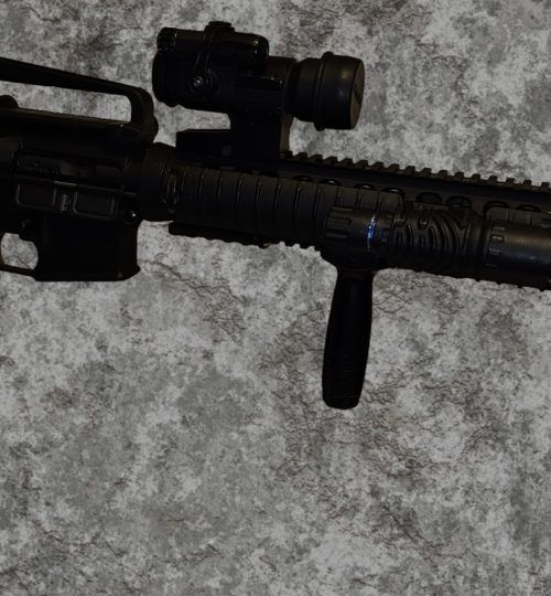 Black rifle with scope and vertical foregrip on a gray carpet.