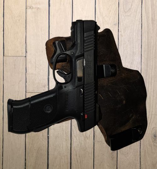 Black handgun resting in a brown holster on a light wood floor.