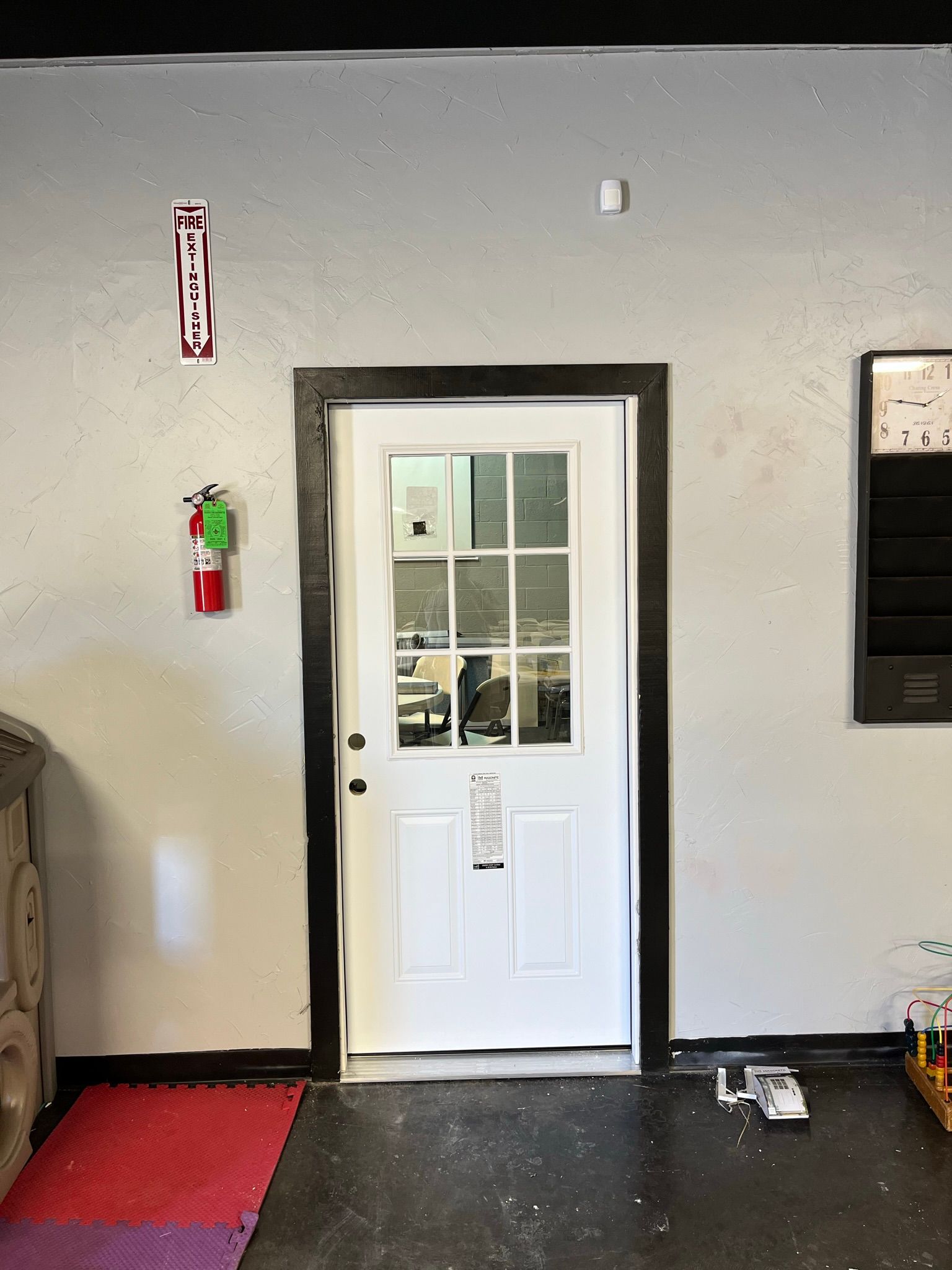 White door with black trim, fire extinguishers, and other items on a gray wall.