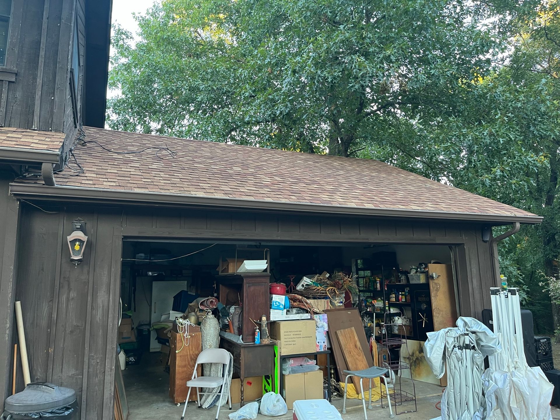 Brown-shingled roof over a cluttered garage. Exterior of a brown house; overgrown trees in background.