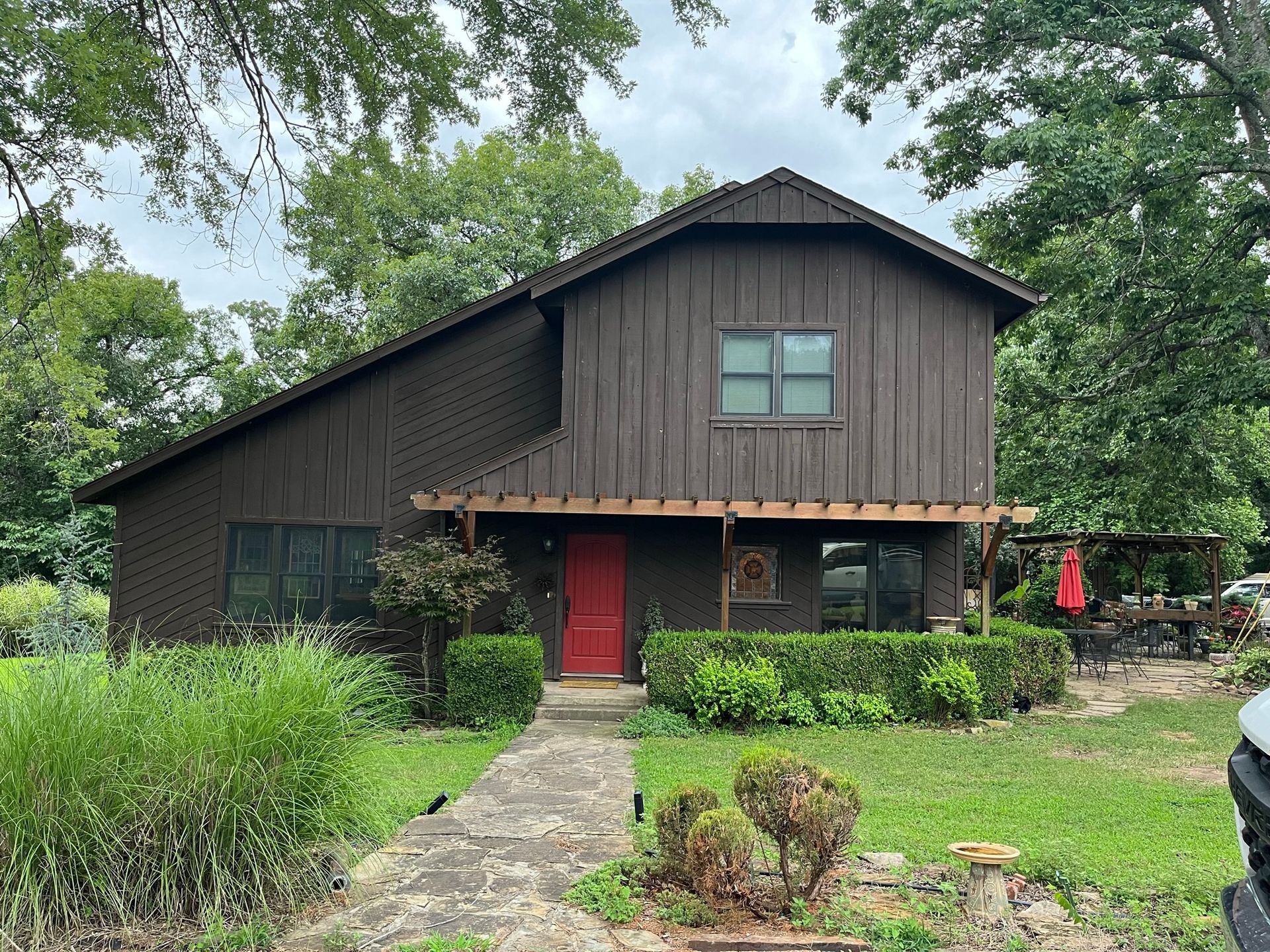 Brown two-story house with red door. Pathway leads to front porch with shrubs. Green grass and trees surround.