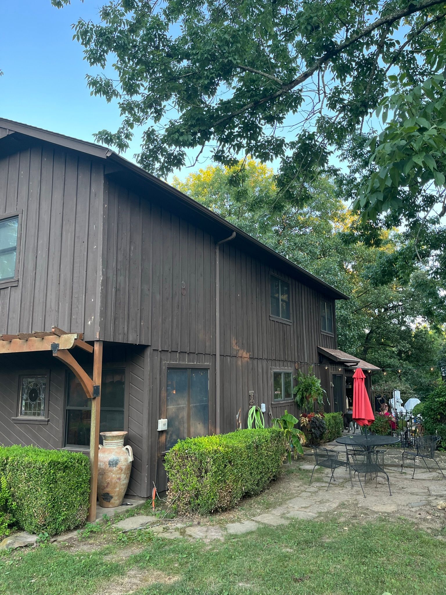 Brown wooden building with dark roof, surrounded by green foliage, outdoor patio with red umbrella.