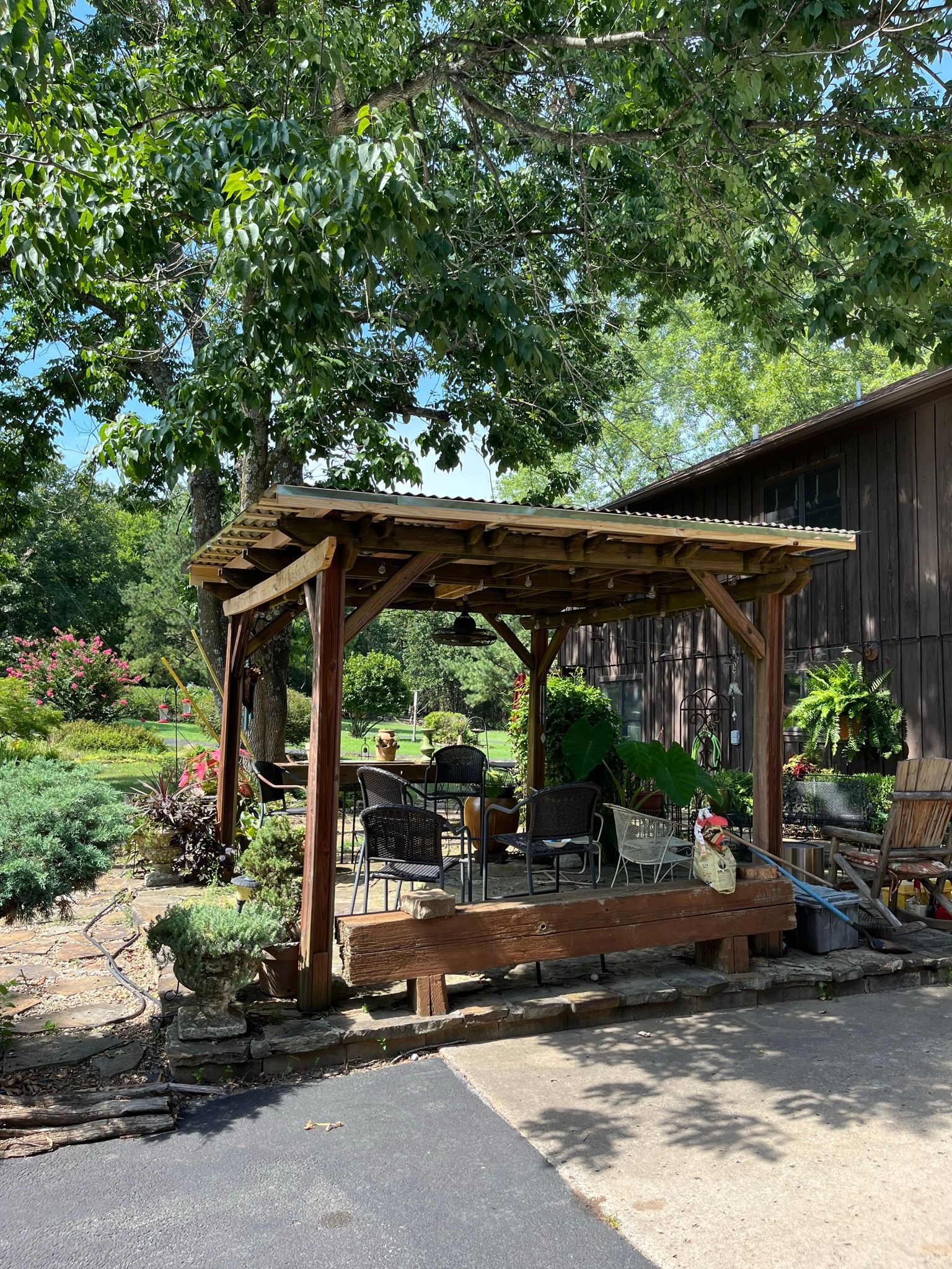 Wooden pergola with seating area; outdoors, next to a building and garden.