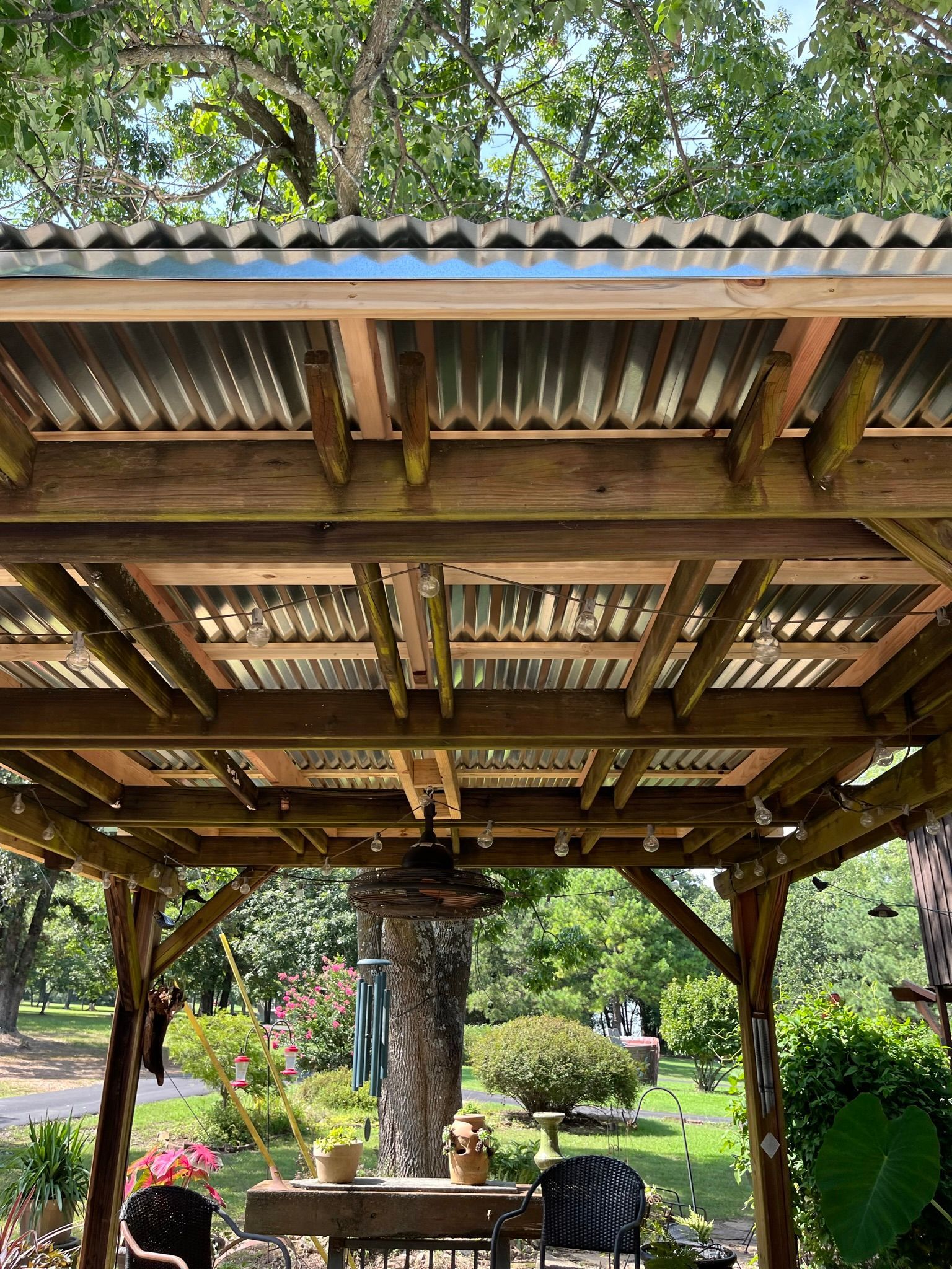 Wooden patio roof with corrugated metal, overlooking a yard with trees and flowers.