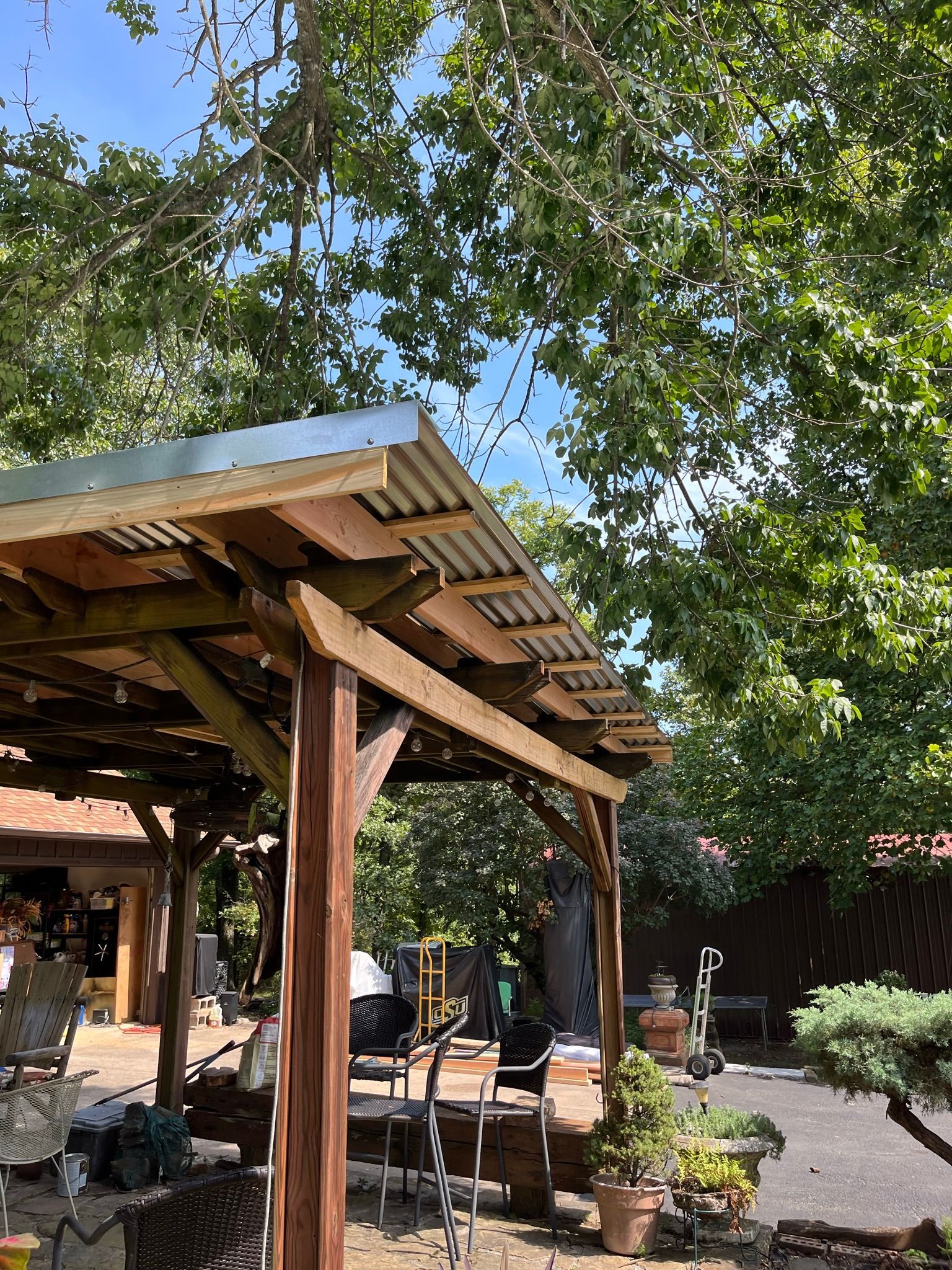 Wooden pergola with corrugated metal roof under a leafy tree, with patio furniture visible.